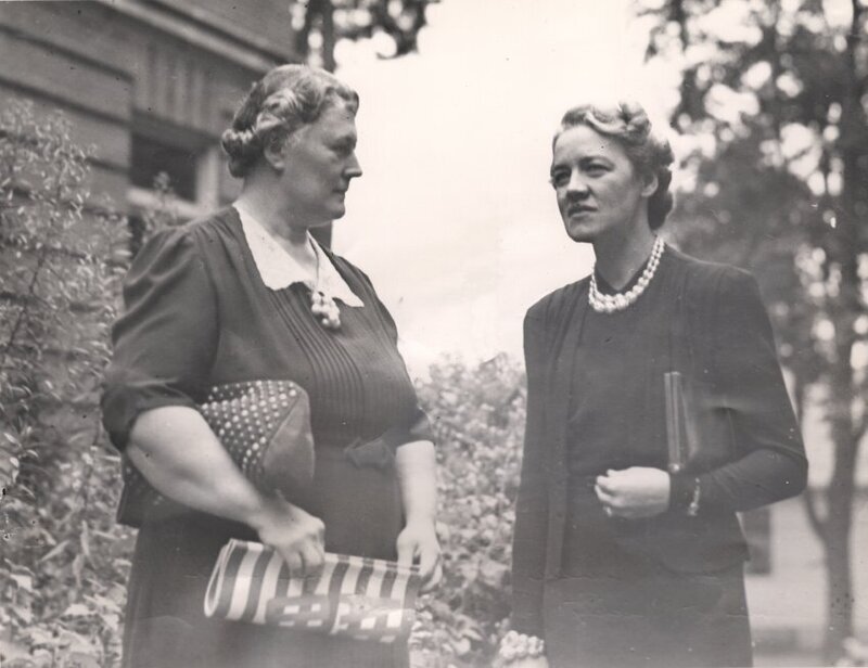 Image of Bertha Bolduc Chaffers, leader of the Women's Division of the Civil Defense Corps in Lewiston-Auburn during WWII, and Senator Margaret Chase Smith. Bertha, a grey haired, heavyset woman wearing a dark colored dress, is standing on the left of the image, looking towards Margaret and holding two purses. Margaret, also grey haired, is standing on the right of the image, facing the camera but looking into the middle distance. Margaret is holidng one purse.