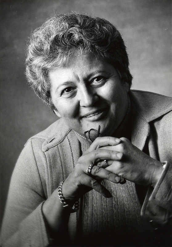 Posed professional portrait of Madeleine Giguere by Tom Jones. The image shows Madeleine, an older woman with grey hair, smiling at the camera while holding her glasses in her hands in front of her chest.